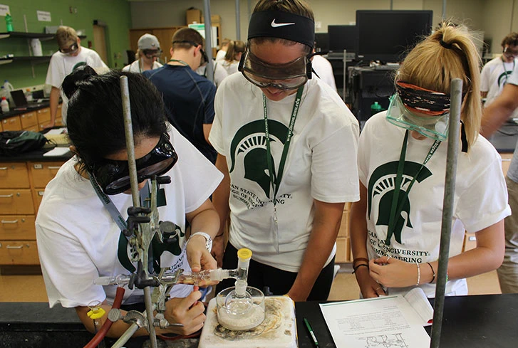 Three diverse female students attending the summer program, hovering over a beaker in a chemical lab