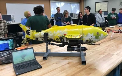 close up of a mechanical yellow fish that is being shown at one of the Engineering preview days. Future students and current MSU students are talking in the background