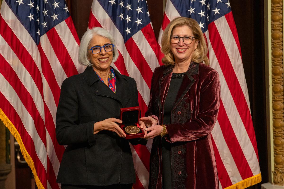 Two women standing in front of American Flags holding medal