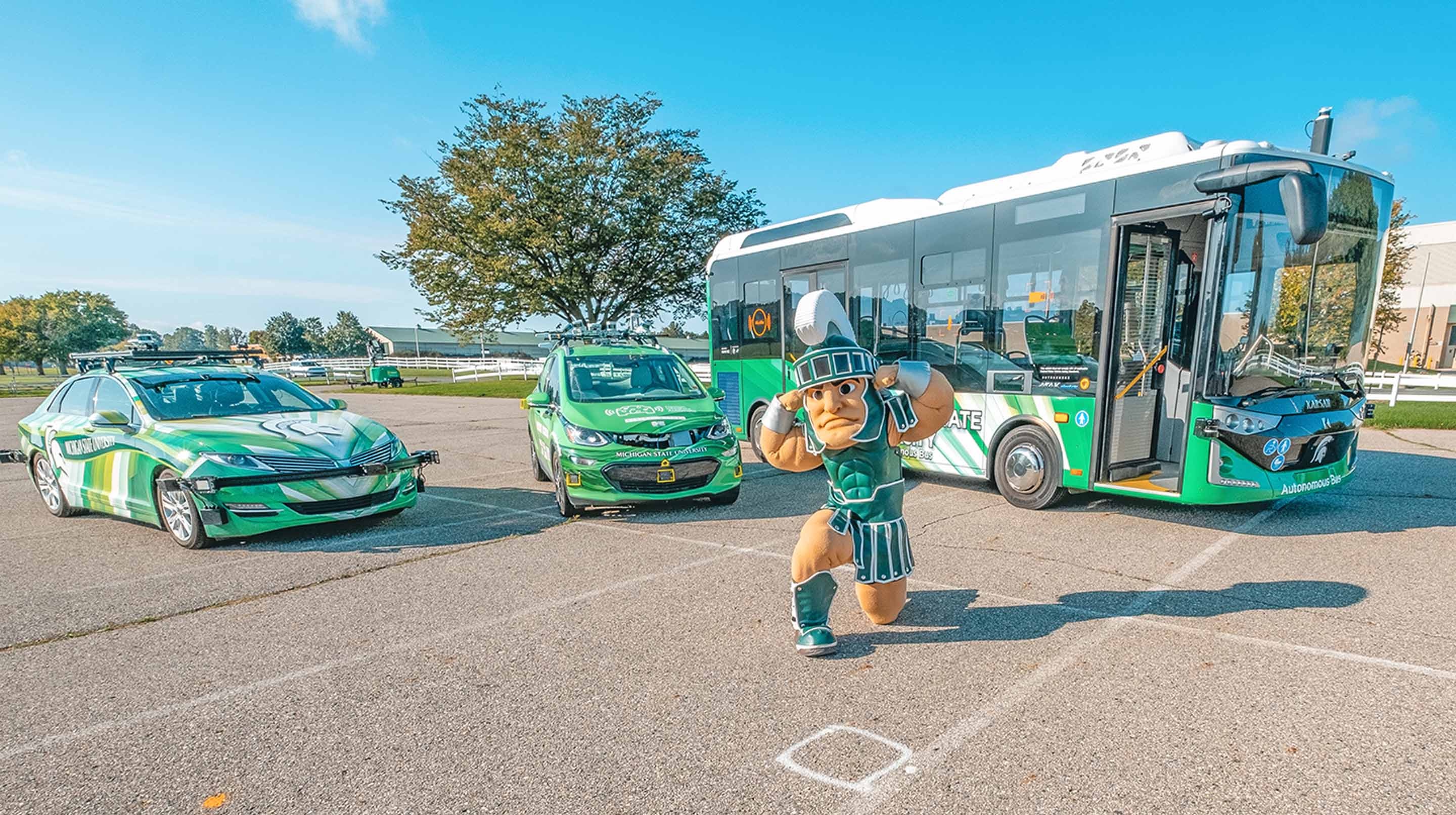The Sparty Mascot poses in front of two cars and a bus, all three green with MSU imagery