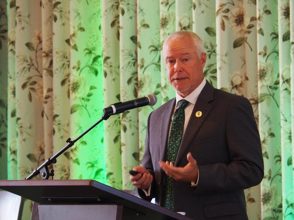 Brad Wieferich in a dark suit and tie, standing in front of a curtain, speaks at a podium.