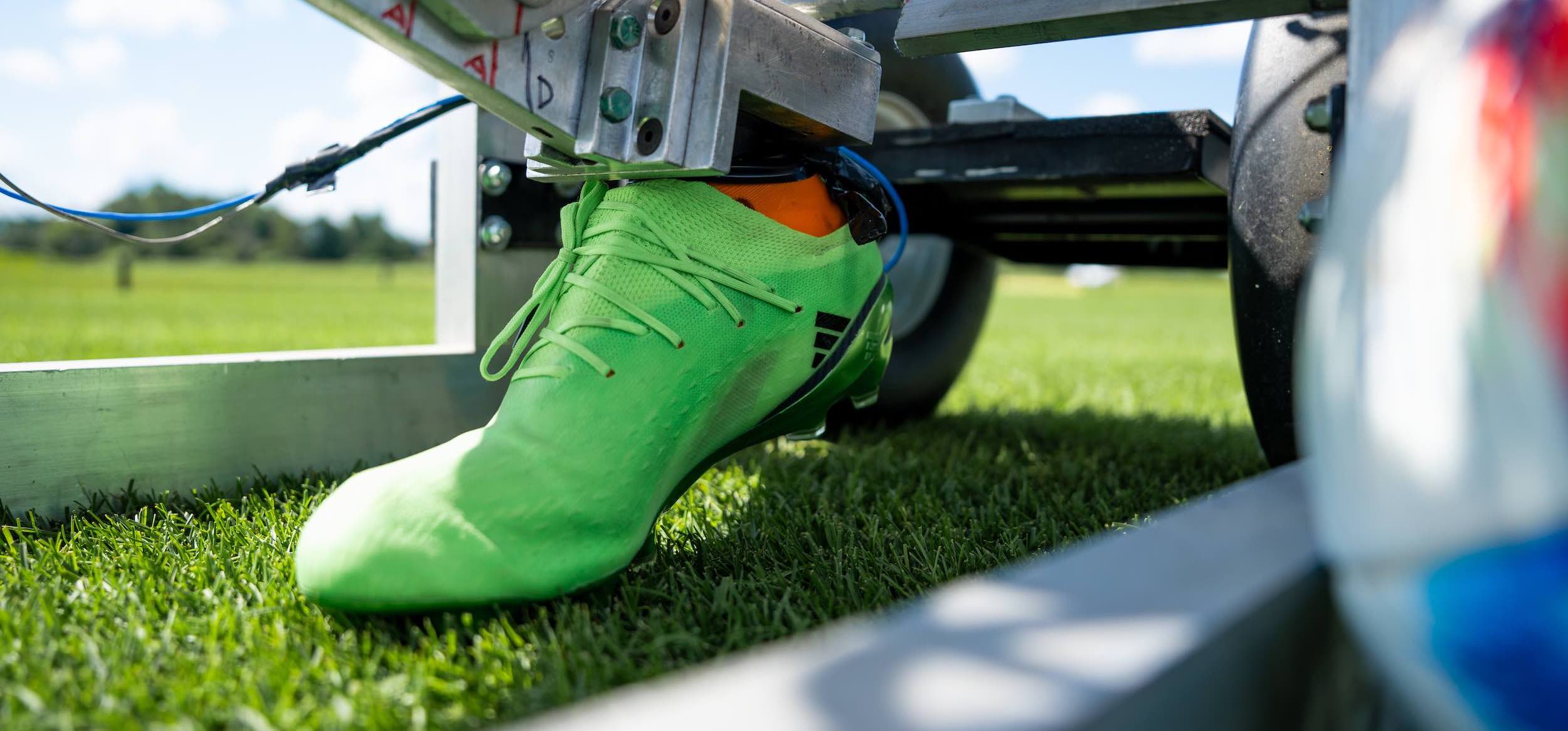A green cleat in steel equipment that moves the cleat like a foot next to grass and an out of focus soccer ball — the equipment and shoe tests the durability of the turf