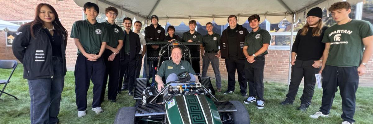 Dean John Papapolymerou sits in a Michigan State Formula Racing car surrounded by student team members during the 2026 Engineering Colloquium.