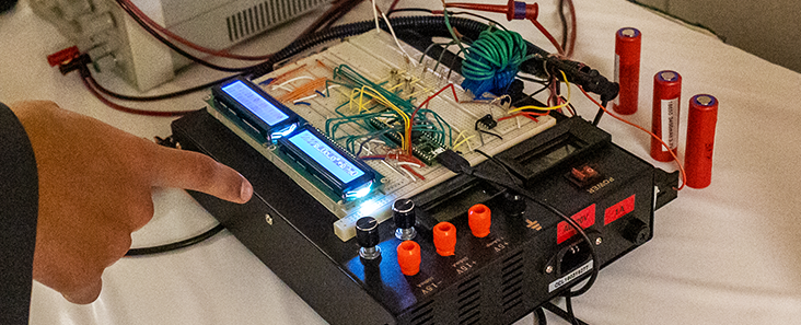 Student pointing at their capstone project, the project is a machine with batteries