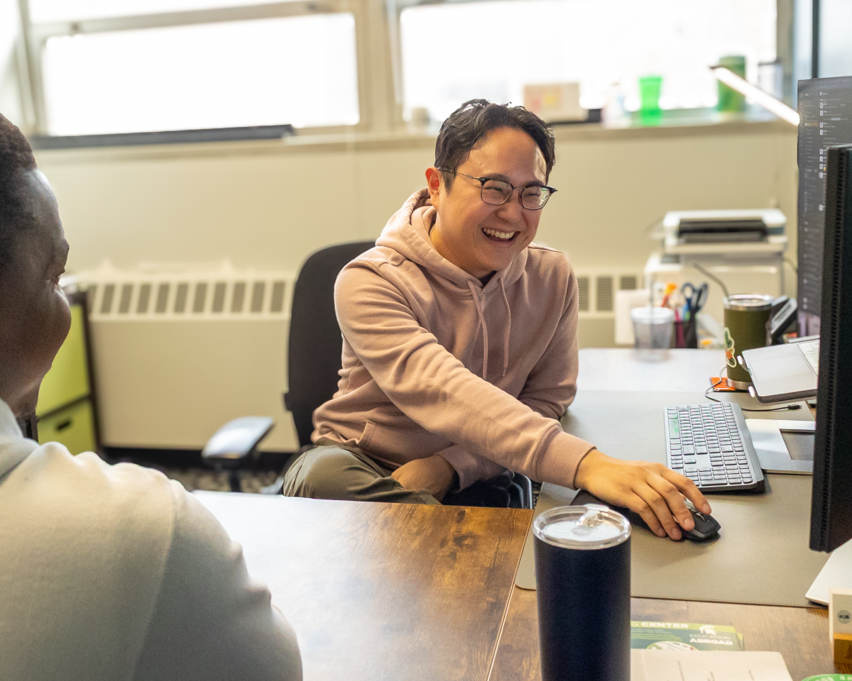 Jeffrey Tsang, smiling, in a beige hoodie, reaches across a desk to move a mouse.