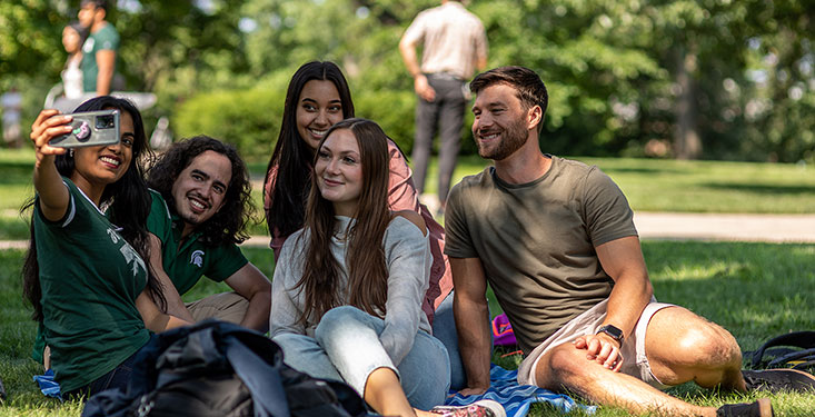 Graduate students sitting on the ground on campus, huddled together to take a picture on one of their phones