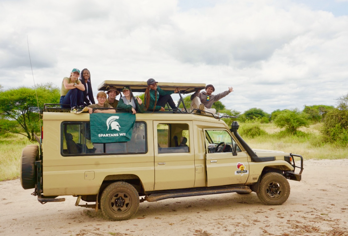 A group of students poses with a Spartans Will flag on top of a safari vehicle in an open savannah.