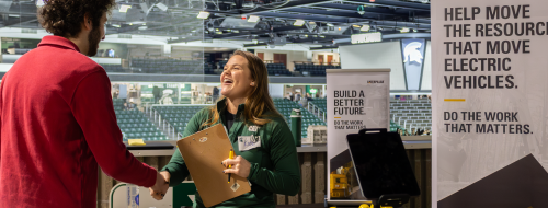 A recruiter at a booth laughs while shaking hands with an attendee at a career fair.